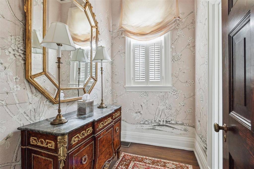 The vanity space of the downstairs powder room exudes elegance with hand-painted floral wallpaper, a gilded mirror, and soft natural light filtering through plantation shutters.
