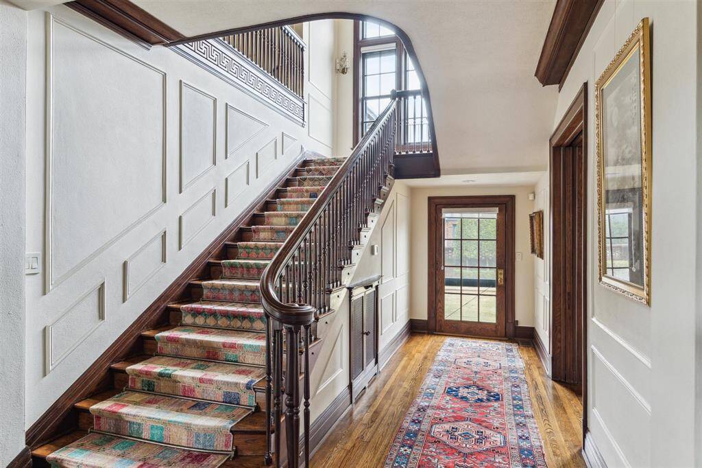 The grand staircase serves as the centerpiece of the home, featuring rich wooden banisters and detailed paneling that exudes timeless elegance. The large windows at the landing fill the space with natural light. The wide, welcoming hallway showcases gleaming hardwood floors and leads to a glass-paneled back door, which offers a seamless connection to the outdoors.
