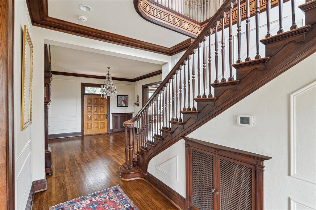 From this angle, the grand staircase with intricate wood and Greek key detailing becomes the focal point of the inviting foyer, which features rich hardwood floors and elegant paneling. The warm tones of the front door and railing are complemented by a sparkling crystal chandelier, adding sophistication. Subtle details like a decorative radiator cover and carved stair balusters enhance the timeless charm of this entryway.