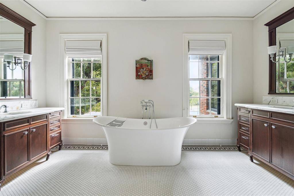The primary bathroom features a luxurious freestanding tub, dual vanities with rich wood cabinetry, large windows that fill the space with natural light, and a separate glass-enclosed shower, creating a serene and elegant retreat.