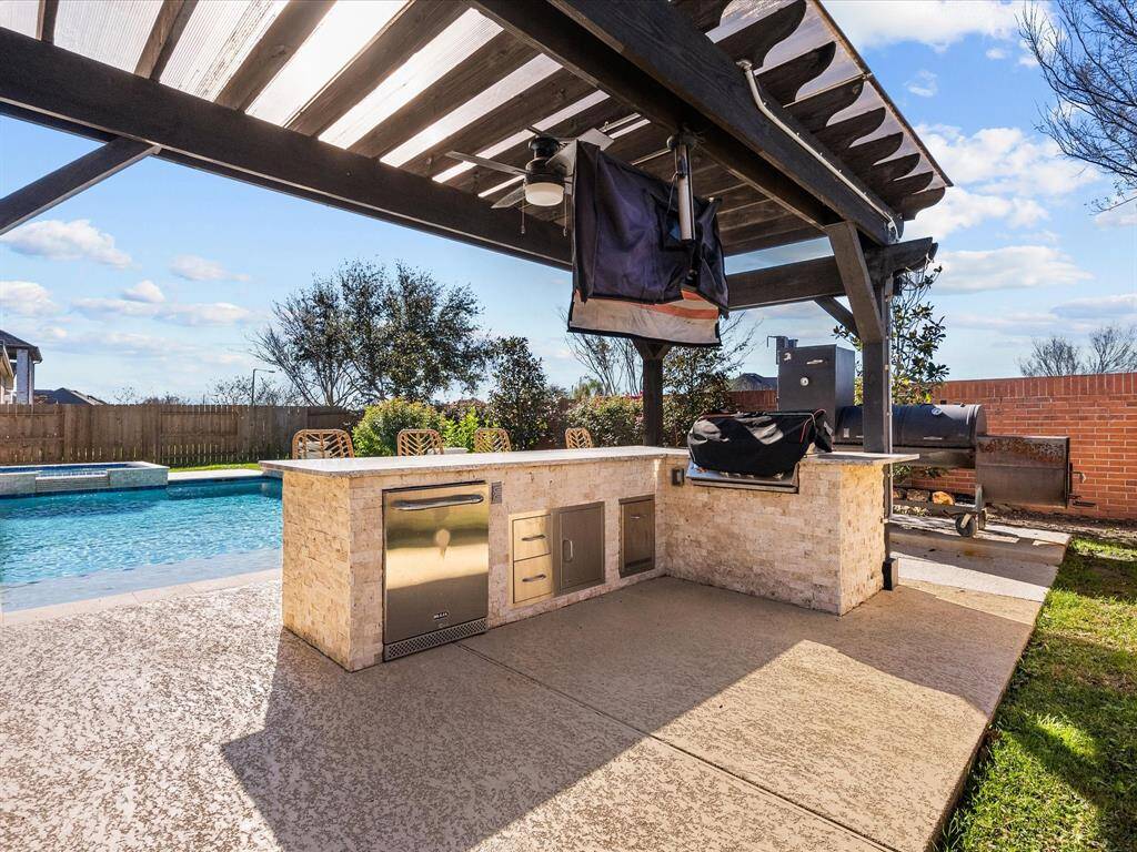 The outdoor kitchen features a built-in BBQ pit, refrigerator, and ample counter space, perfect for poolside entertaining. The pergola provides shade and a stylish focal point.