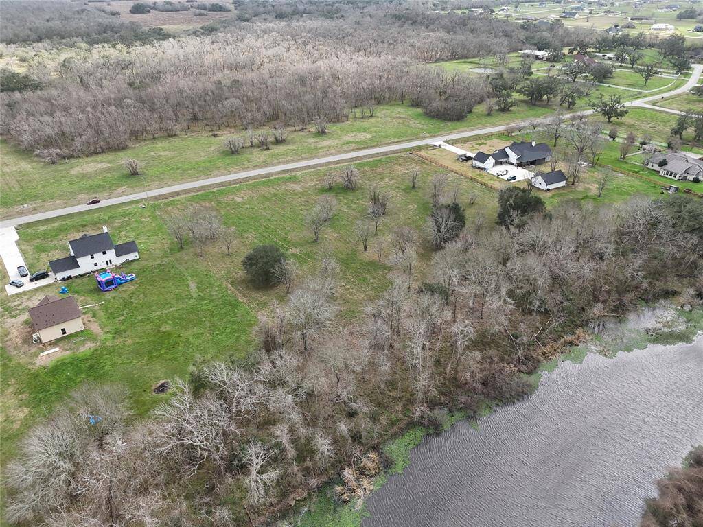 This photo shows a spacious property, surrounded by open fields and a wooded area. A small road runs through the area, and there's a water feature at the edge, offering a peaceful, scenic setting.