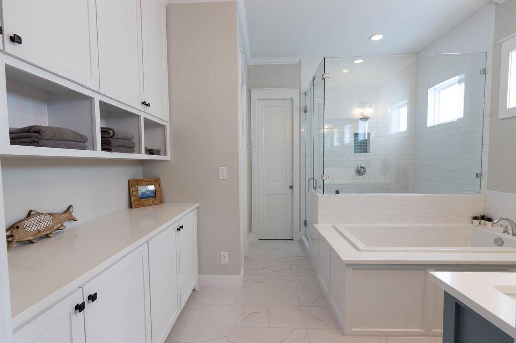 This bathroom features a sleek, modern design with white cabinetry and open shelving. It includes a spacious bathtub and a glass-enclosed shower. Natural light filters through small windows, creating a bright and inviting space.