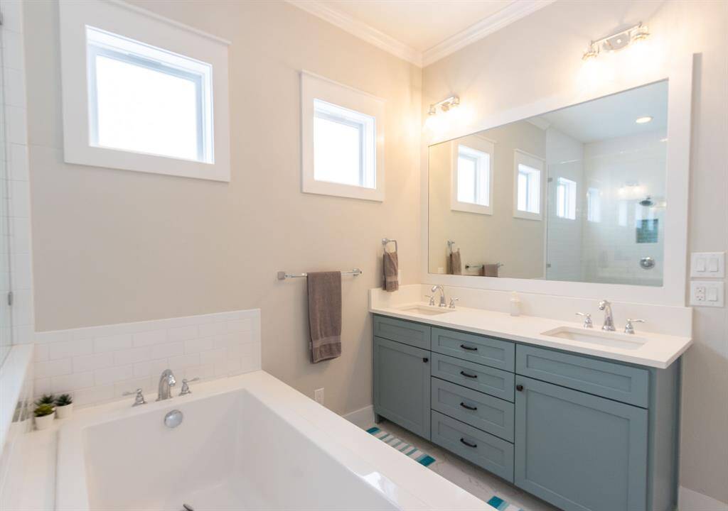 This bathroom features a spacious soaking tub, dual sinks with modern fixtures, and ample storage with stylish cabinetry. It has a clean, bright design with natural light from three windows, complemented by elegant tile flooring