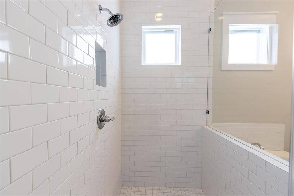 This modern bathroom features a spacious walk-in glass shower and a large soaking tub. It has bright tile work, ample natural light from the high windows, and a neutral color palette, creating a clean and inviting space.