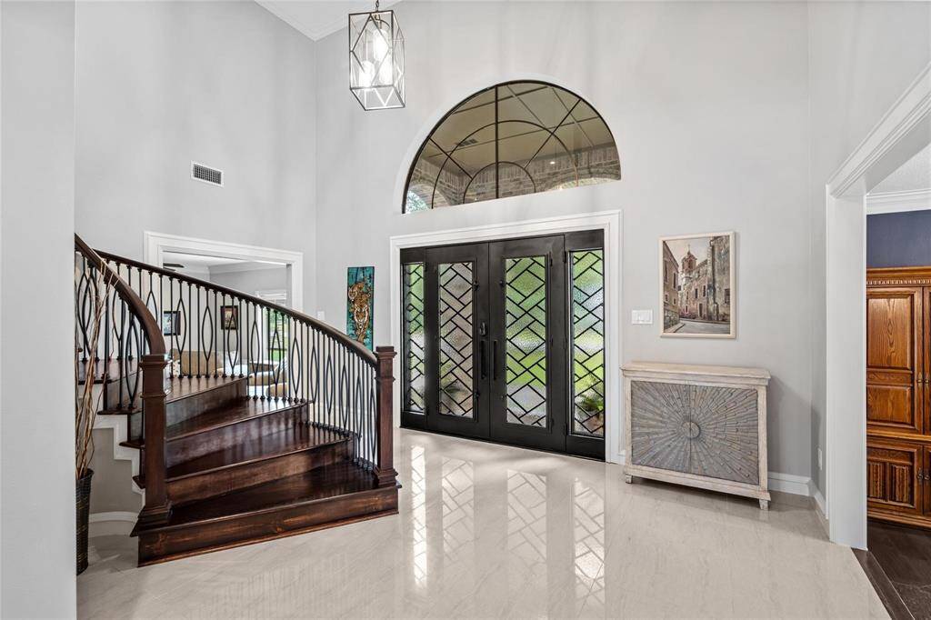 This elegant entryway features a grand curved staircase with dark wood steps and wrought iron railings. The large, decorative glass double doors allow natural light to flood in, complementing the high ceilings and modern chandelier. Neutral walls and polished floors create a welcoming, sophisticated atmosphere.