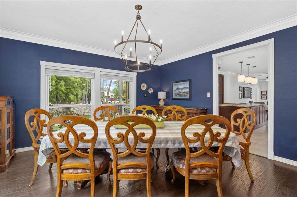 This dining room features a bold accent wall, a large wooden table with elegant chairs, and a classic chandelier. Natural light pours in through the windows, offering a view of lush greenery. The space is open and connects seamlessly to the kitchen area.