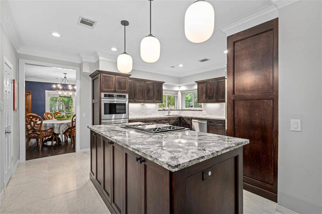 This kitchen features elegant dark wood cabinetry, a large marble island with a built-in gas cooktop, and modern pendant lighting. It opens to a dining area with a classic chandelier, creating a seamless flow for entertaining.