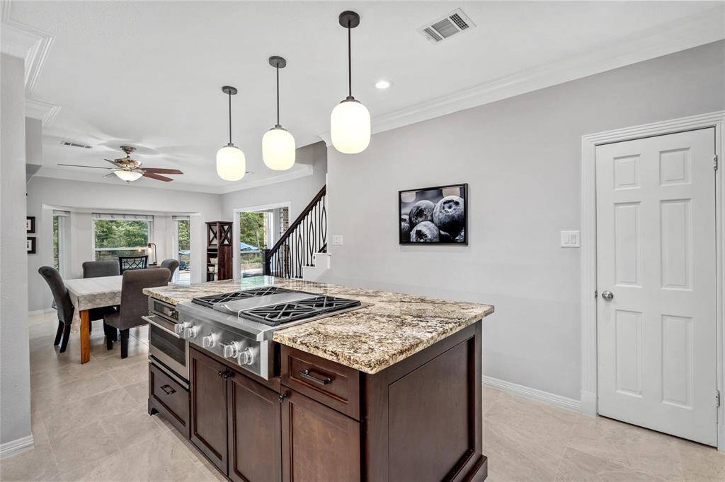 This photo showcases a modern kitchen with a central island featuring a gas stove and granite countertops. The space is well-lit with pendant lights and opens to a dining area with large windows and a view of the staircase. Neutral tones and sleek finishes create a welcoming atmosphere.