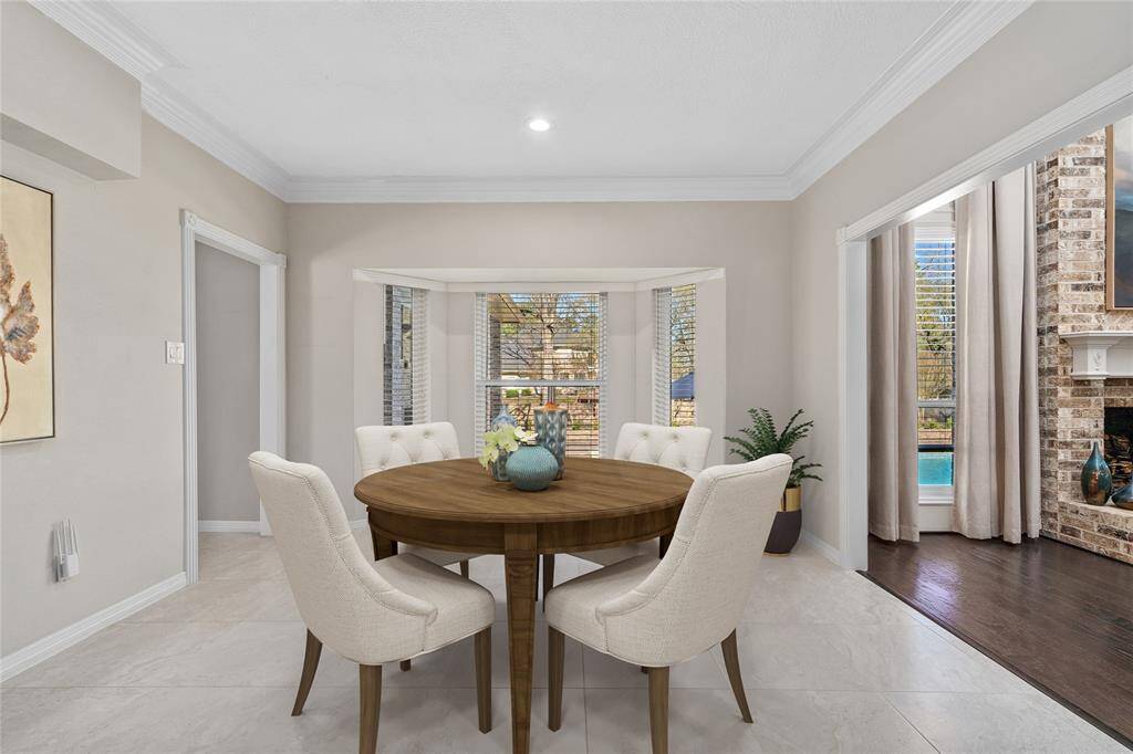 This inviting dining area features a round wooden table, upholstered chairs, and large windows that flood the space with natural light. Adjacent is a cozy living area with a charming brick fireplace.