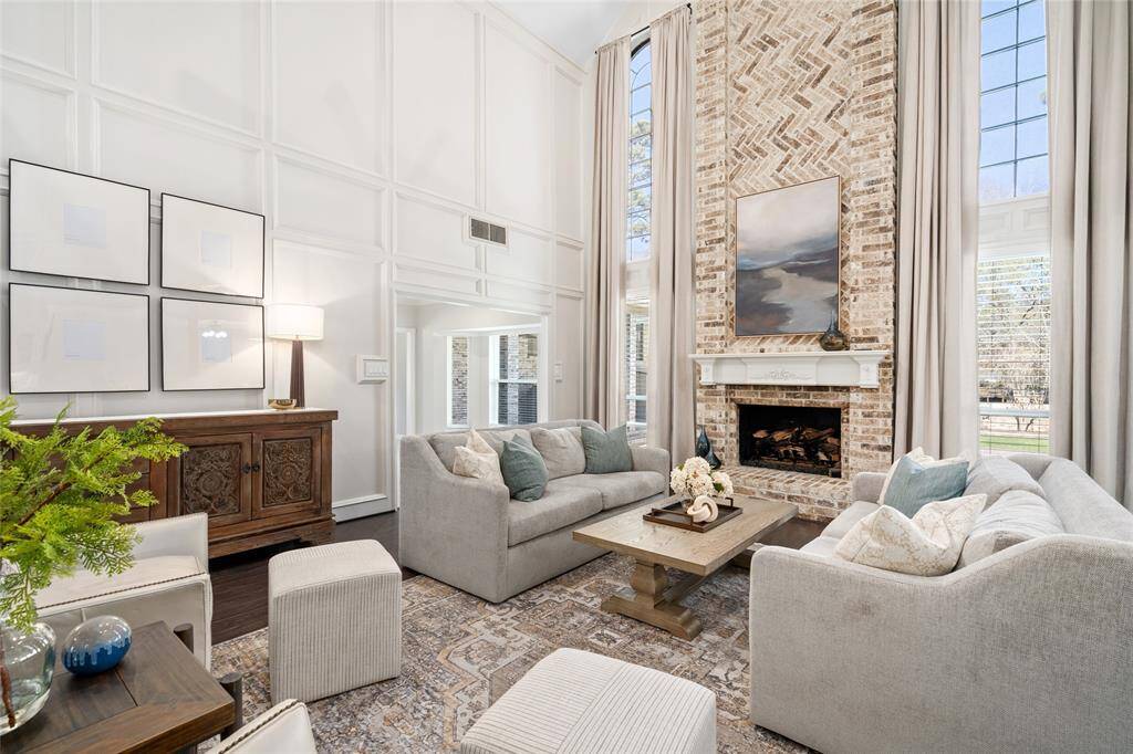 This inviting living room features high ceilings, a floor-to-ceiling brick fireplace, and large windows that flood the space with natural light. The neutral-toned decor and plush seating create a cozy yet elegant ambiance, perfect for family gatherings.
