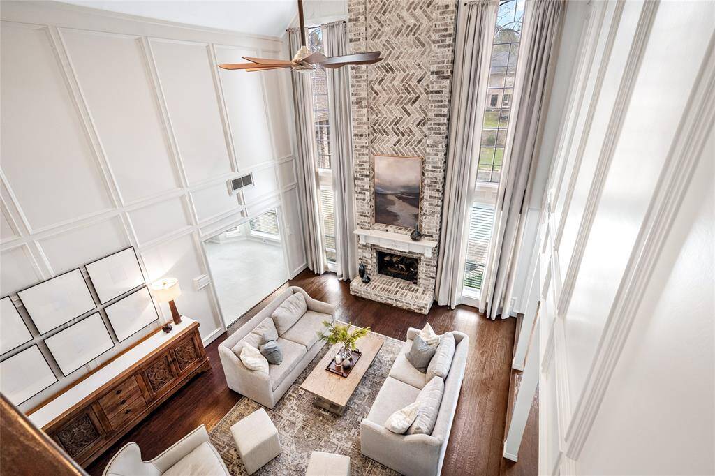 This spacious living room features high ceilings, a stylish brick fireplace, and large windows for natural light. The elegant decor includes a neutral color palette, comfortable seating, and a fan for added comfort, creating a welcoming and airy atmosphere.