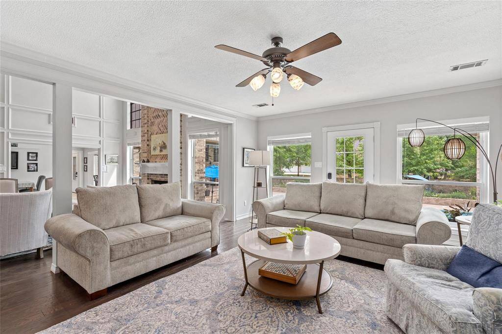 Bright and inviting living room with neutral-toned sofas, a stylish ceiling fan, and ample natural light from large windows. Open layout connects to a dining area, creating a spacious and cozy atmosphere.