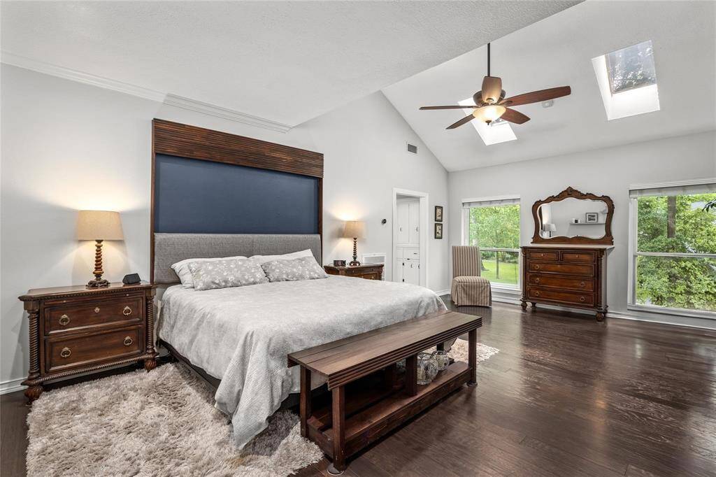 This spacious bedroom features a cozy bed with elegant wooden furniture, including a dresser and nightstands. The room is enhanced by natural light from large windows and skylights, highlighting the rich hardwood floors and high ceiling with a stylish ceiling fan.