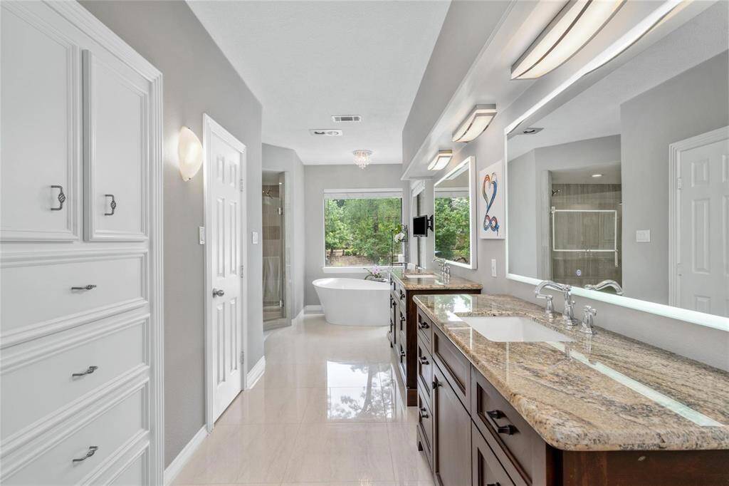 This spacious bathroom features dual sinks with granite countertops, ample cabinetry, and a large window. A freestanding bathtub and a separate glass-enclosed shower provide luxury and relaxation, while light neutral tones create a bright and inviting atmosphere.