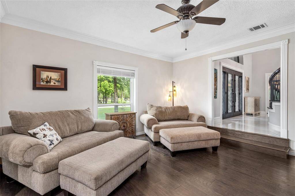 Cozy living room with warm wood flooring, comfortable seating, and a ceiling fan. Large window provides natural light and views of lush greenery. An open doorway leads to a bright entryway with elegant double doors and a staircase.