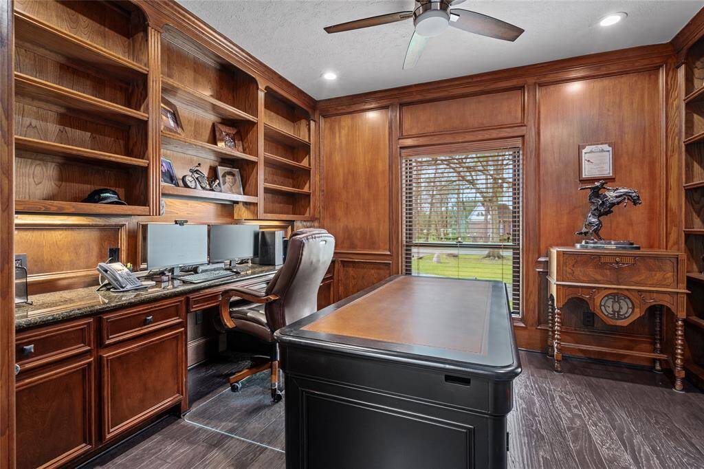 This is a warm, elegant home office with rich wooden built-in shelves and cabinetry. It features a sleek desk, dual monitors, and a large window offering a view of the outdoors, creating an inviting and productive workspace.