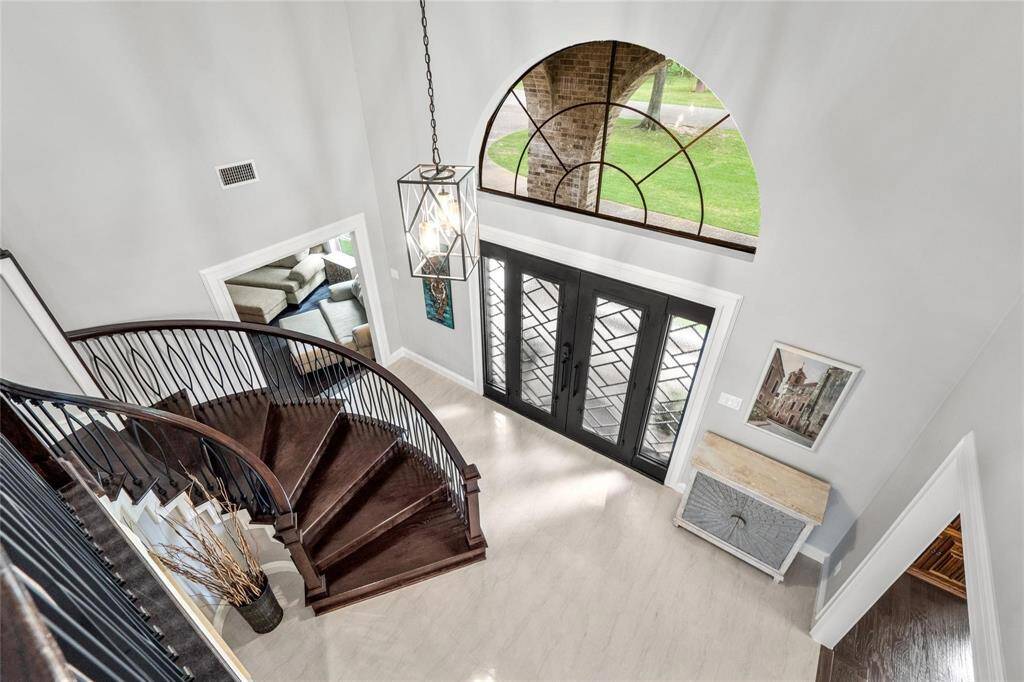 This elegant entryway features a grand, curved staircase with dark wood steps and wrought iron railing, leading to a well-lit foyer. The space is enhanced by large double doors with decorative glass, a high arched window, and stylish light fixture, offering a welcoming and sophisticated first impression.