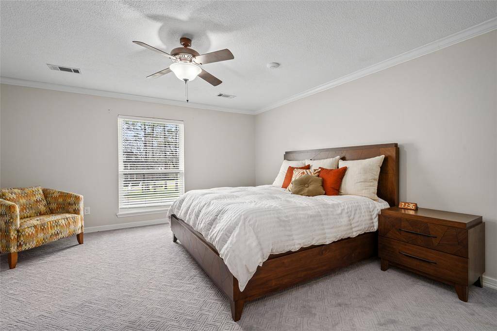 This bedroom is spacious and bright, featuring neutral walls and carpet. It includes a large window, modern ceiling fan, and a cozy bed with vibrant accent pillows. A stylish armchair and wooden nightstands add warmth and functionality to the room.
