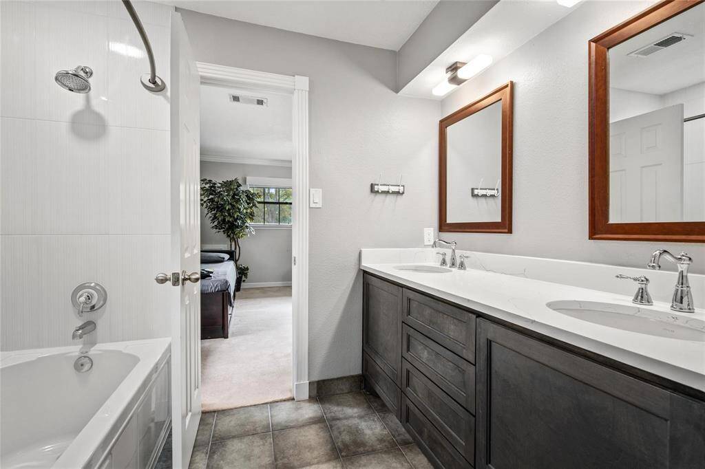 This bathroom features a modern dual-sink vanity with sleek fixtures, a large mirror, and a bathtub with a shower. The space is bright, with a neutral color palette, and opens to a cozy bedroom with natural light from a window.