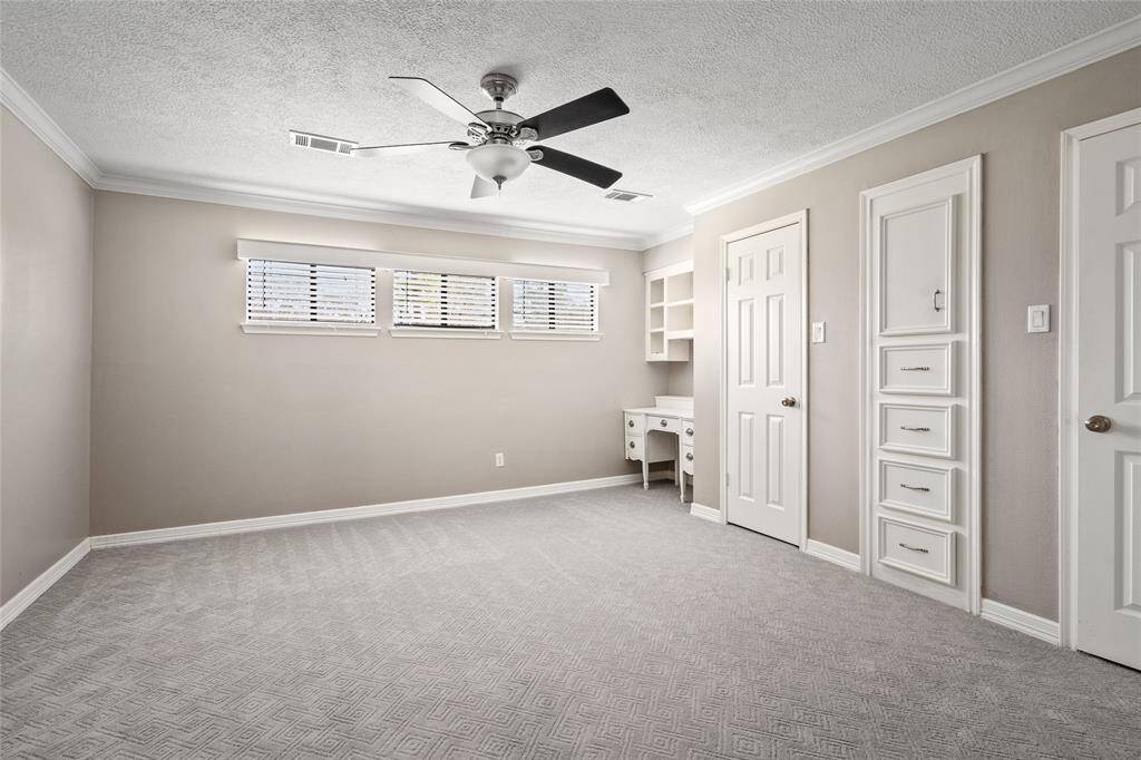 This spacious bedroom features neutral walls, a ceiling fan, and natural light from three windows. It includes built-in shelves, drawers, and a small desk, offering ample storage and workspace.