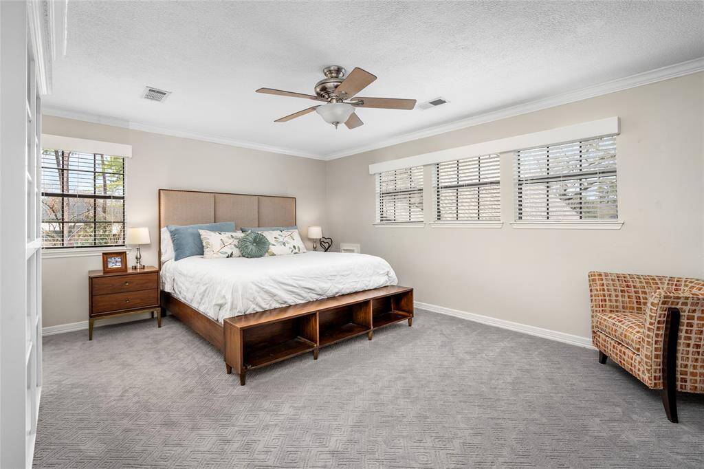 This spacious bedroom features a comfortable bed with storage, large windows with blinds offering ample natural light, a ceiling fan, and neutral-colored walls and carpet. A cozy chair and nightstand complete the inviting setup.