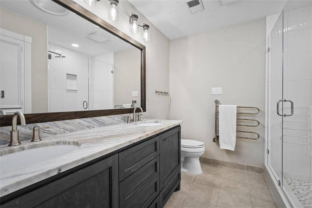 This bathroom features a modern design with a spacious double vanity topped with marble, a large mirror, and elegant light fixtures. It includes a glass-enclosed shower and neutral tile flooring, offering a clean and stylish look.