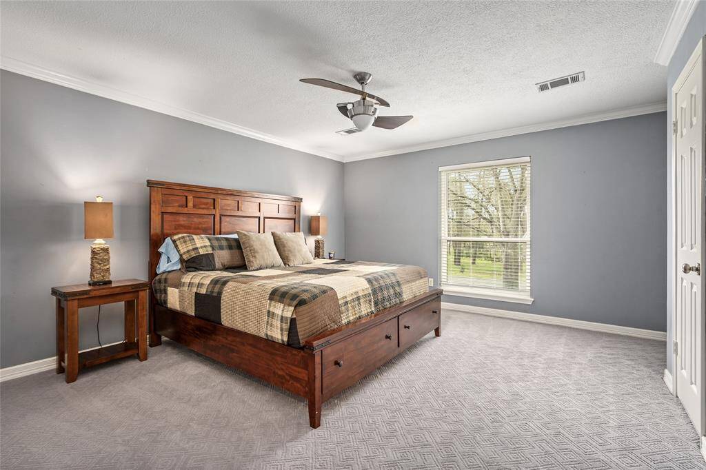 This spacious bedroom features a large window with a scenic view, a ceiling fan, and cozy carpeting. The room is tastefully decorated with a wooden bed frame and matching nightstands, creating a warm and inviting atmosphere.
