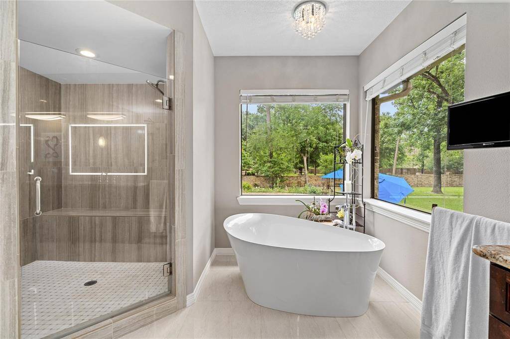 This elegant bathroom features a spacious glass shower and a sleek standalone tub, with large windows showcasing a lush backyard view. Neutral tones and modern lighting enhance its serene, luxurious feel.
