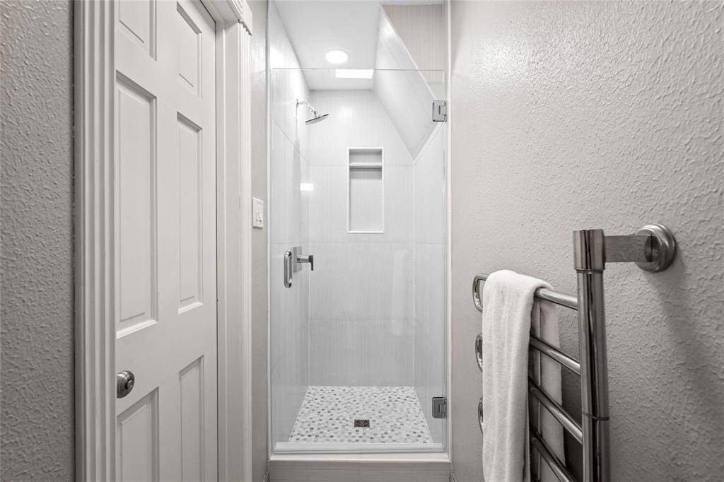 A updated shower with a glass door, featuring a small built-in shelf and light gray tiling. The space is well-lit with recessed lighting, and there's a sleek towel rack on the wall.