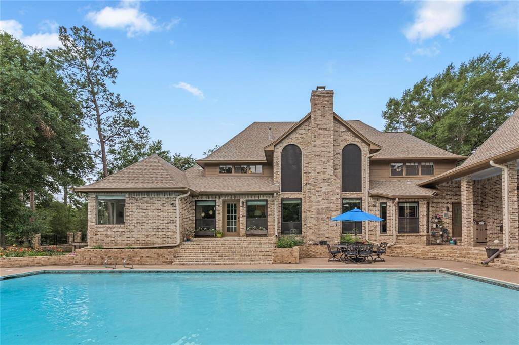 This photo showcases a spacious brick home with a large swimming pool in the foreground. The house features multiple gables, tall windows, and a prominent chimney. There is a patio area with outdoor seating and an umbrella, surrounded by mature trees, offering a serene and private setting.