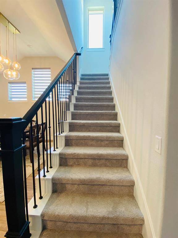 This photo features a carpeted staircase with black iron railings leading to a bright upper level. Natural light streams in from a tall window at the top, and adjacent modern lighting fixtures enhance the open, airy feel.
