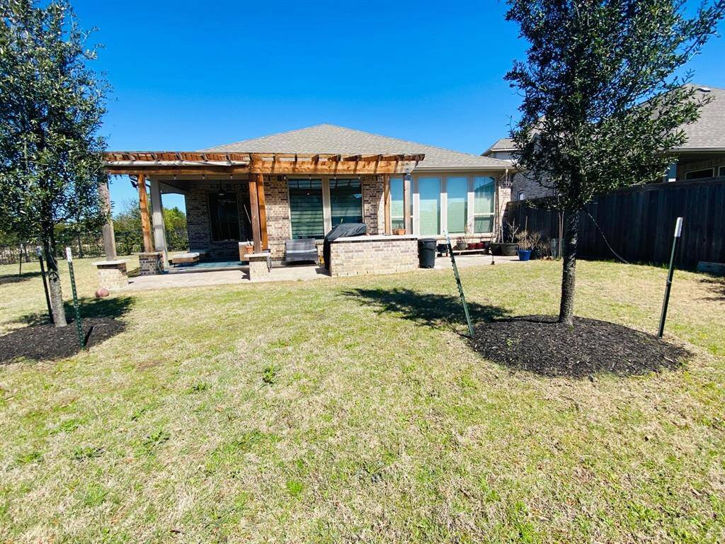 This photo shows a spacious backyard with a well-maintained lawn and two young trees. The house features a large patio area with a pergola, stone accents, and ample seating space, perfect for outdoor gatherings.
