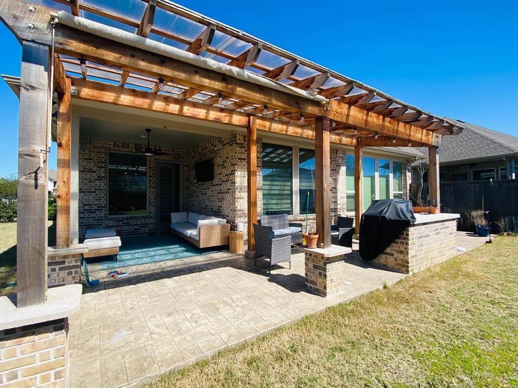 This photo showcases a charming backyard patio with a wooden pergola, comfortable seating, a TV, and a built-in grill area. The space is perfect for outdoor entertaining and relaxation, set against a backdrop of a well-maintained lawn.