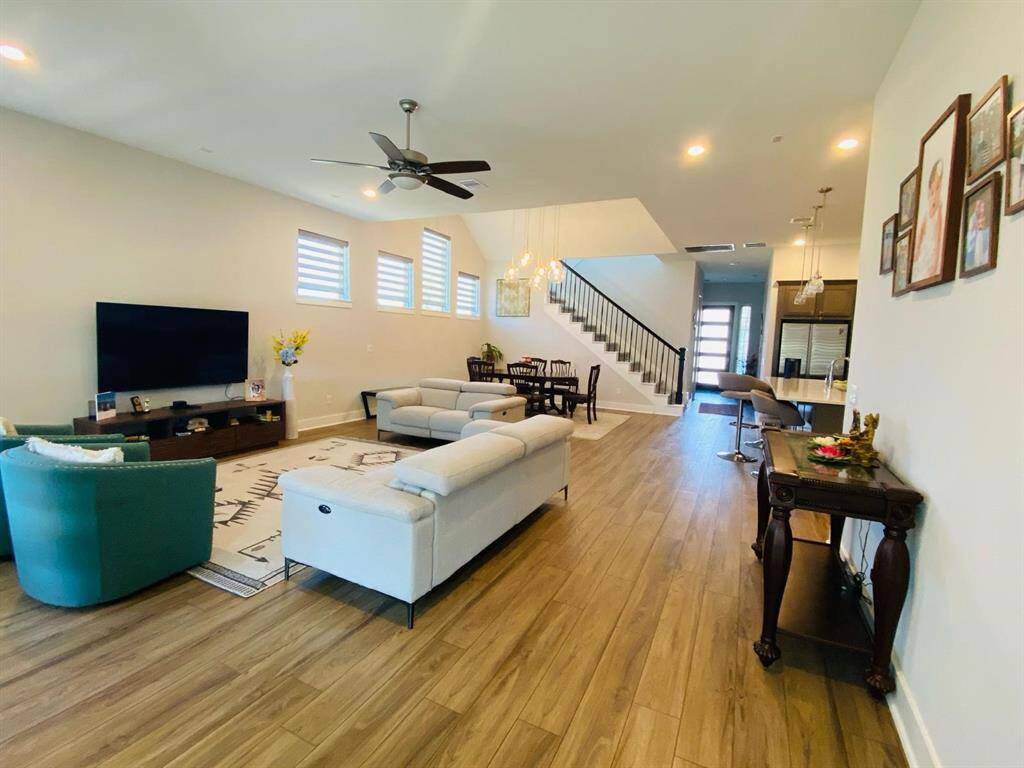This spacious, open-concept living area features wood flooring, a cozy seating arrangement, and modern decor. It seamlessly connects to a dining area and kitchen with bar seating. The room is well-lit with large windows, and a staircase leads to the upper floor.