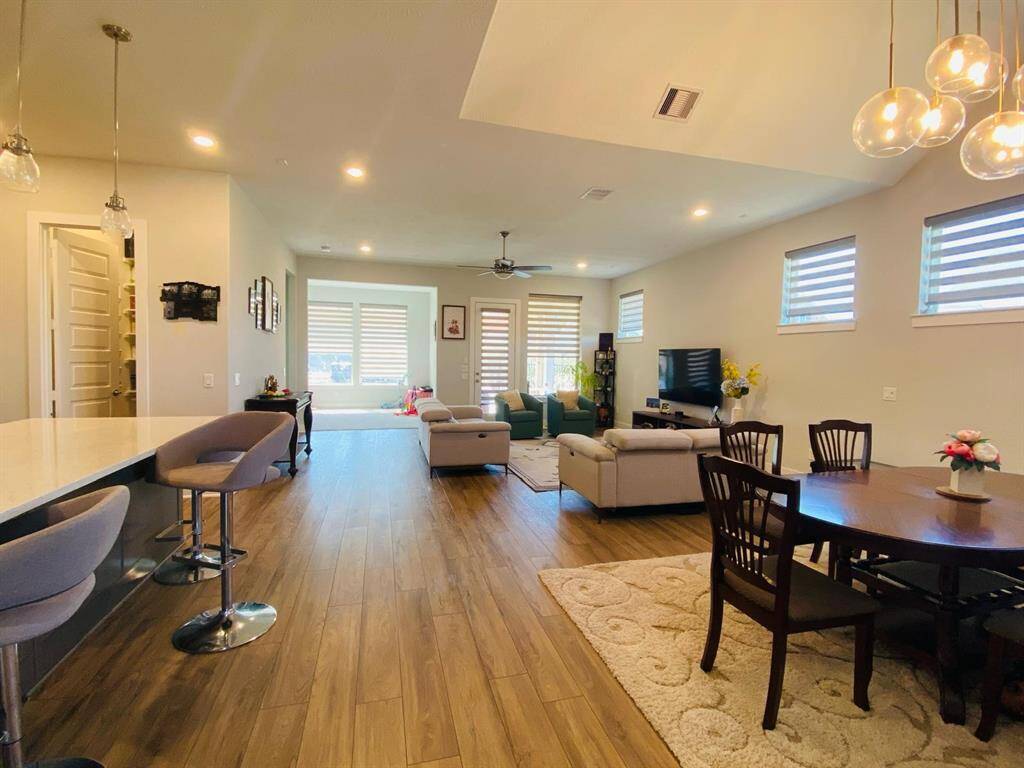 This spacious open-concept living area features modern wood flooring, a cozy sitting area with ample natural light, a stylish dining space, and a sleek kitchen island with bar seating. Perfect for family gatherings and entertaining.