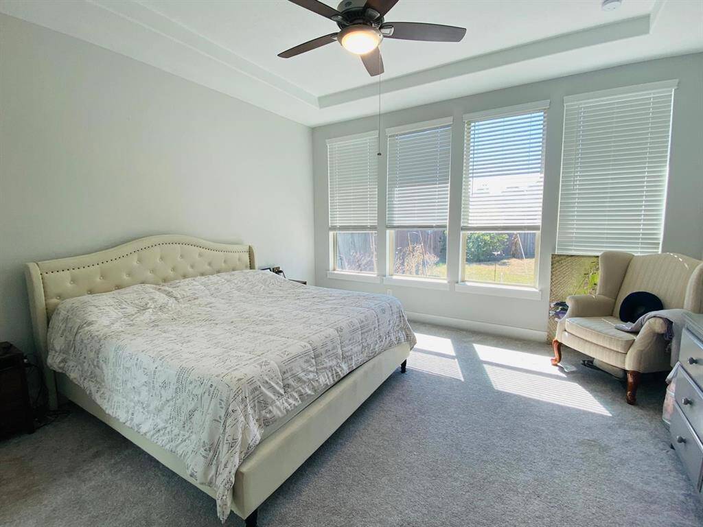 This spacious bedroom features large windows that fill the room with natural light, a ceiling fan, and a cozy bed. It has soft carpeting and a comfortable armchair, creating a relaxing atmosphere.