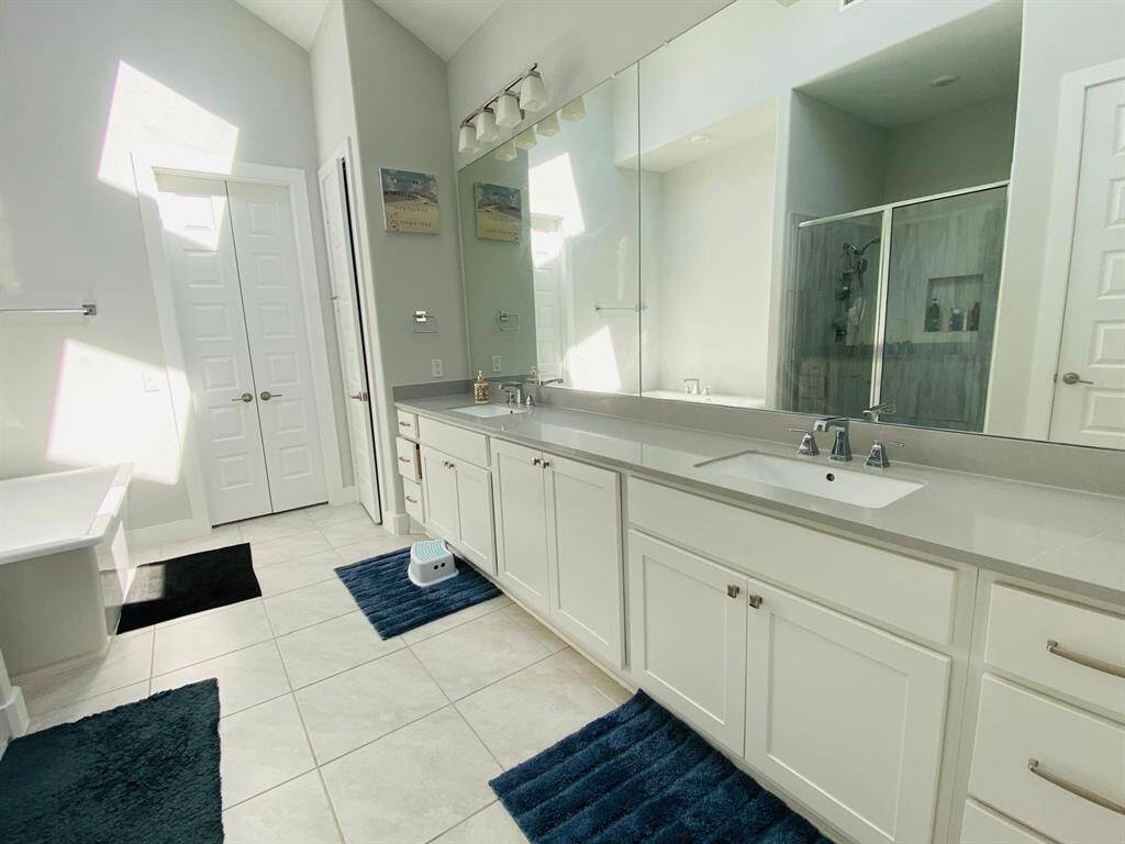 This is a spacious, modern bathroom featuring a double vanity with ample counter space, large mirrors, and white cabinetry. It has a glass-enclosed shower, a bathtub, and bright natural lighting, creating a fresh, inviting atmosphere.