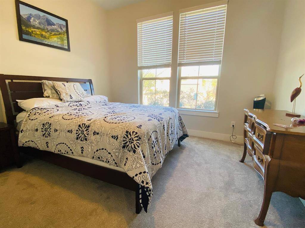 This cozy bedroom features large windows for natural light, neutral walls, and carpeting. It includes a comfortable bed with a decorative bedspread and a classic wooden dresser, creating a warm and inviting atmosphere.
