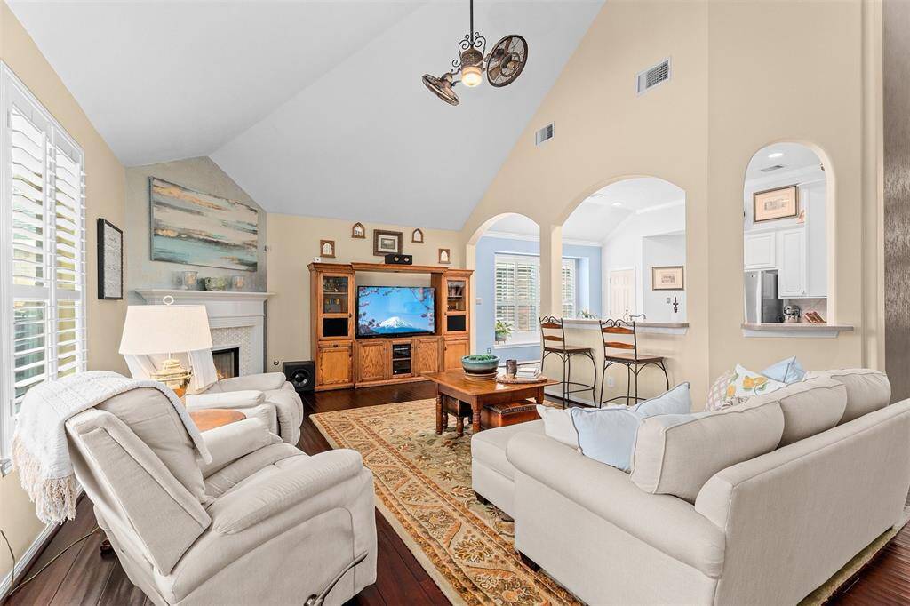 This inviting living room features high vaulted ceilings, a cozy fireplace, and abundant natural light. It flows seamlessly into the adjacent kitchen area through open archways.