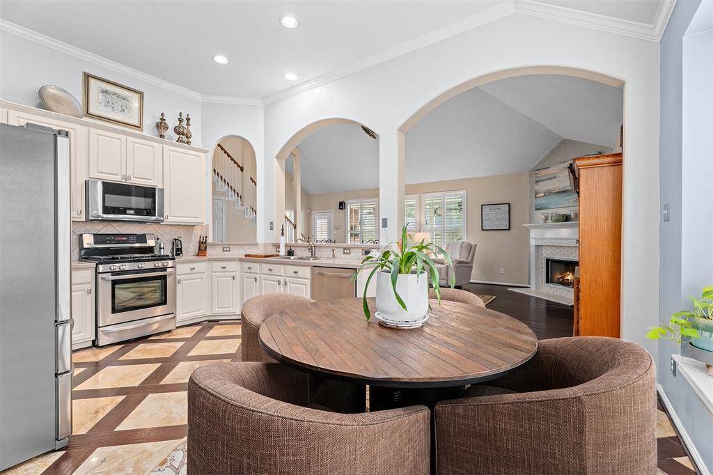 This cozy dining area features a round table with comfortable chairs, set against a large window with plantation shutters. The space seamlessly connects to an open kitchen with stainless steel appliances and a living room with a fireplace, creating a warm and inviting atmosphere.