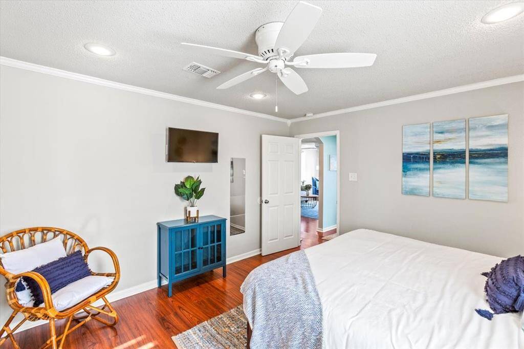 Bright bedroom featuring polished hardwood floors, a modern ceiling fan, recessed lighting, and contemporary decor. It includes a mounted flat-screen TV, a wicker chair, and a blue cabinet, with a serene ocean-themed triptych on the wall.