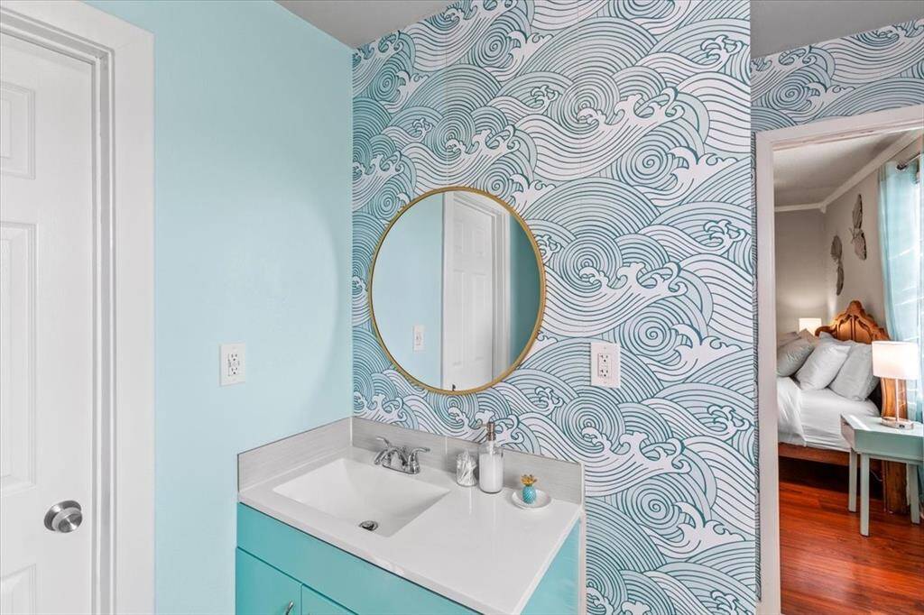 This is a bright bathroom with a light blue color scheme, featuring a distinctive wave-patterned wallpaper accent wall and a modern vanity with a coordinating blue cabinet. A circular mirror adds a contemporary touch, and the room opens into a bedroom area, suggesting an en suite layout.