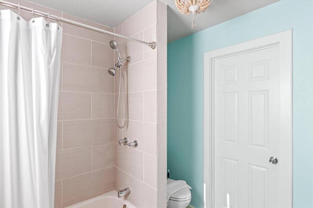 This is a modern bathroom featuring a combination tub and shower with a dual showerhead system, light beige tile walls, and a bright teal accent wall. The space includes a white door and a simple white curtain, creating a clean and refreshing look.