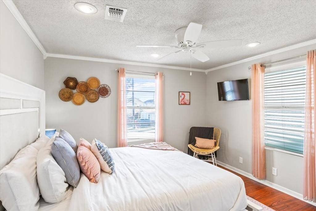 This is a cozy, well-lit bedroom featuring a large bed, hardwood floors, ceiling fan, decorative wicker wall accents, and a mounted TV. Large windows with blinds provide ample natural light, complemented by soft, peach-colored curtains.