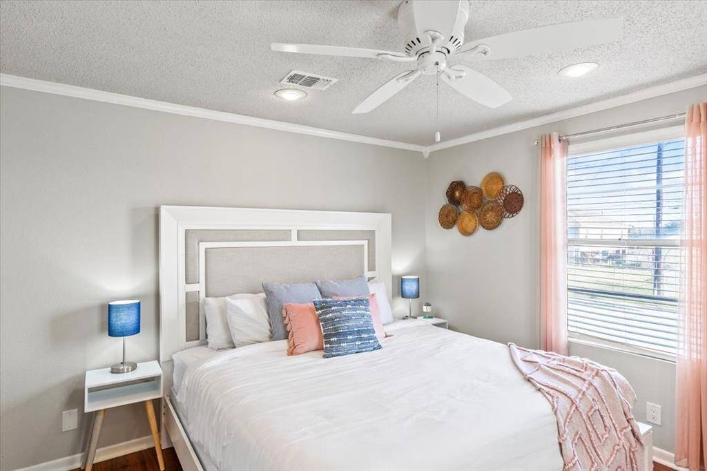 This is a cozy, well-lit bedroom featuring a large bed with a stylish headboard, two bedside tables with lamps, a ceiling fan, and a window allowing for natural light, adorned with pink curtains. Decorative wall baskets add a touch of warmth to the space.