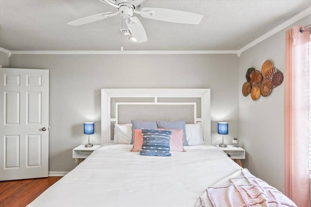 This is a cozy, well-lit bedroom featuring a large bed with a stylish headboard, two bedside tables with lamps, a ceiling fan for comfort, and hardwood flooring. The room is complemented by neutral wall colors and accented with decorative wall baskets and light curtains, creating a warm and inviting space.