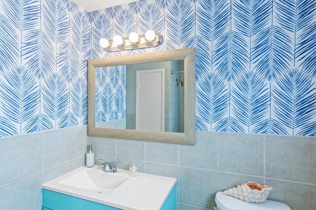 This is a bright, modern bathroom featuring an eye-catching blue and white palm leaf-patterned wallpaper, a large mirror with a textured frame, a white countertop with a built-in sink, and a vibrant turquoise vanity. The space is complemented by soft grey tiles and a well-lit vanity area. Hall full bath. One of 2 bathrooms on main level.