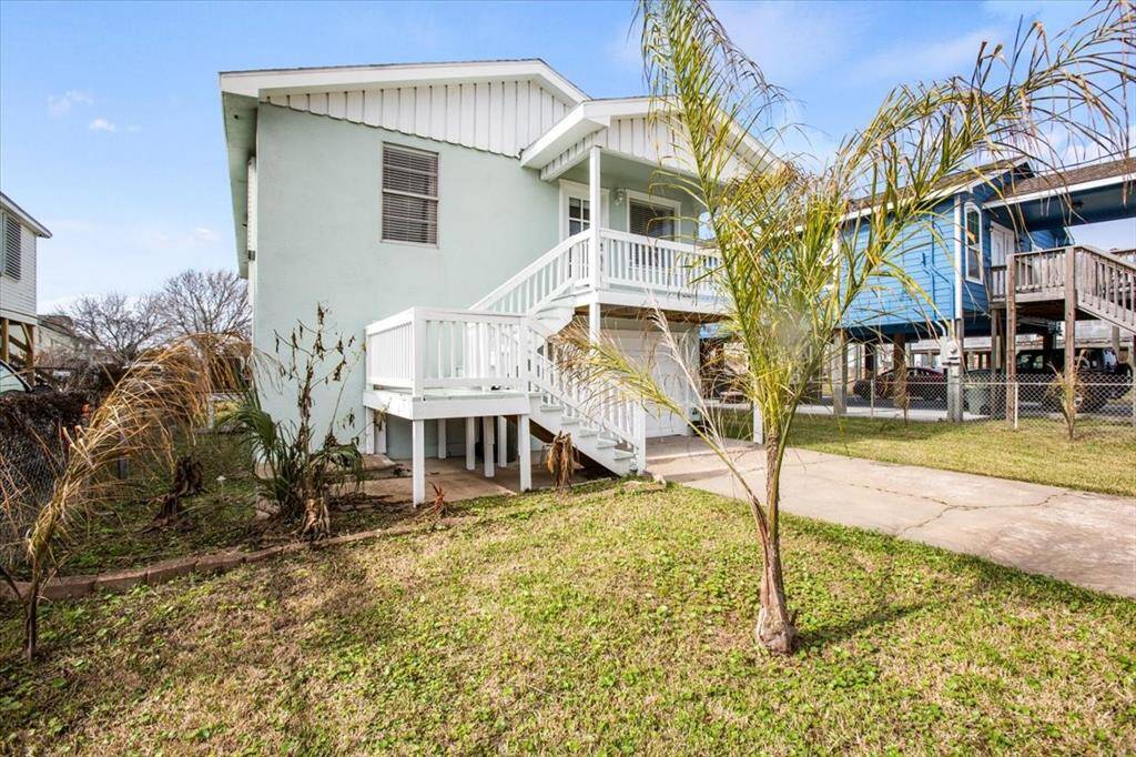 This is a two-story, elevated, light-colored home featuring an external staircase leading to the upper floor, a balcony, and a small front yard with some foliage. The house is situated in a neighborhood with similar elevated homes.