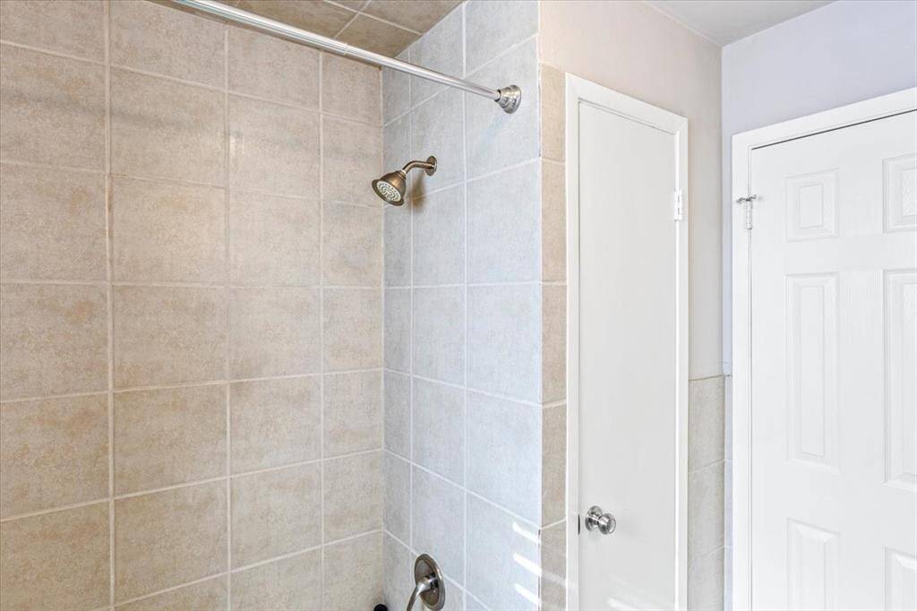 This is a clean, well-maintained bathroom with neutral-toned wall tiles, featuring a shower area with a wall-mounted showerhead and a glass door leading to a white interior door.